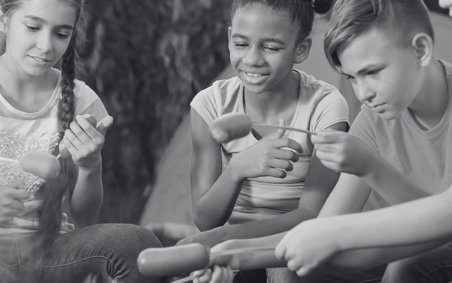Drei Teenager sitzen um ein Lagerfeuer und halten Würstchen an Spießen über das Feuer. Das Bild ist in Schwarz-Weiß