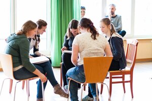Das Bild zeigt eine Gruppe junger Menschen, die in einem hellen Raum mit großen Fenstern zusammenarbeiten. Sie sitzen auf Stühlen im Kreis, haben Notizbücher oder Papier vor sich und wirken konzentriert und engagiert. Im Hintergrund beobachten zwei Erwachsene die Gruppe mit einem freundlichen und interessierten Ausdruck. Die Atmosphäre wirkt entspannt und produktiv, was auf eine kreative oder pädagogische Gruppenaktivität hindeutet.