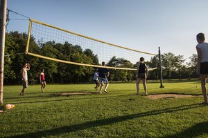 Eine Gruppe von Menschen spielt Beachvolleyball auf einer grünen Wiese unter blauem Himmel. Das Volleyballnetz ist leicht durchhängend gespannt. Die Spieler sind in sommerlicher Kleidung, einige barfuß, und bewegen sich aktiv über das Spielfeld. Im Hintergrund sind Bäume und Natur zu sehen, was eine entspannte und sportliche Atmosphäre vermittelt.