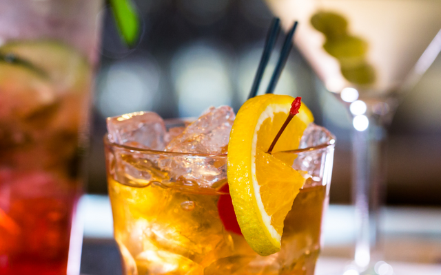 Ein Glas mit einem Cocktail auf Eis, garniert mit einer Orangenscheibe und einer Kirsche, steht im Vordergrund auf einem Tisch. Im unscharfen Hintergrund sind zwei weitere Cocktailgläser zu sehen.