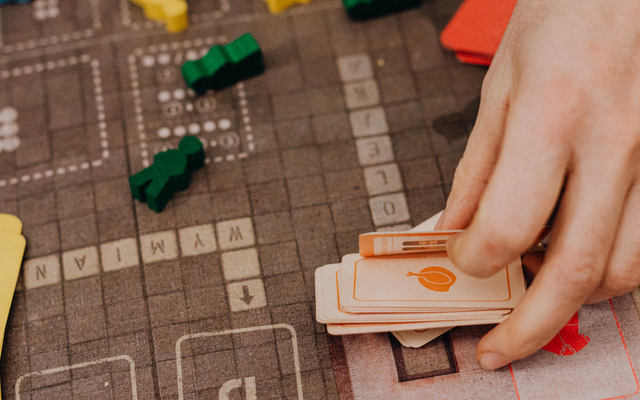 Ich überlege Eine Nahaufnahme zeigt die Hände einer Person, die orangefarbene Spielkarten von einem Spielbrett nimmt, auf dem sich bunte Spielfiguren befinden.